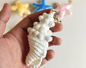 May include: A white crocheted seashell keychain with a textured, spiraled design and a gold-toned metal ring. In the background, three additional crocheted starfish keychains in yellow, blue, and light pink are visible, each with a gold-toned ring.