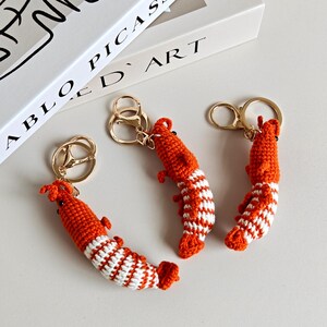 May include: Three crochet shrimp keychains featuring orange and white yarn segments, small black bead eyes, and gold-toned metal split rings. Each shrimp has a curved body shape with textured stitching and small appendages along the sides.