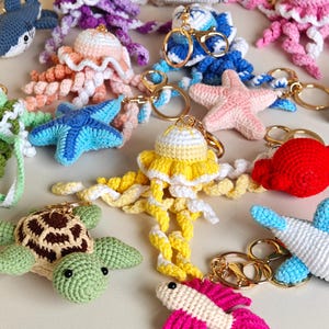 May include: A collection of various crocheted sea creature keychains including jellyfish, starfish, a turtle, and a fish. Each item features a gold-toned metal keyring and clasp. The creatures are crafted in vibrant colors like yellow, blue, pink, red, and green.