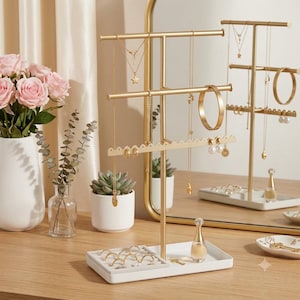 May include: A gold-toned metal jewelry stand with three horizontal bars and a white rectangular base. The stand features a scalloped edge for earrings, hooks for necklaces, and a smooth bar for bracelets. It is displayed on a wooden surface.