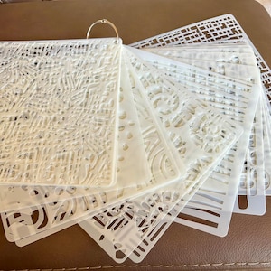 May include: A set of translucent white plastic stencils held together by a metal ring. Each rectangular stencil features a unique laser-cut pattern, including geometric grids, organic swirls, and intricate line designs for various crafting projects.