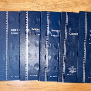 May include: A set of seven blue Whitman coin collection folders featuring embossed circular indentations for coin storage. The covers display white text including Indian Head, Buffalo, Mercury, Roosevelt, and Washington Quarter Collection titles.