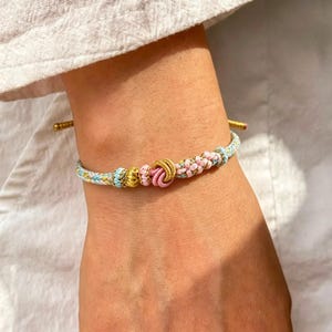 Puede incluir: Una pulsera de cordón trenzado con un nudo central rosa, flanqueado por secciones trenzadas en dorado, azul claro y rosa. La pulsera incluye extremos de cordón ajustables y muestra un patrón multicolor de hilos en rosa pastel, azul claro y dorado.