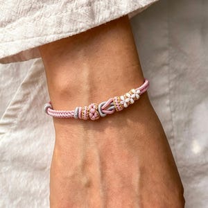 May include: A woven cord bracelet featuring a light pink base with accents of metallic gold, silver, and white thread. The design includes a central knot detail and a series of small, white, flower-shaped embellishments along the side of the band.