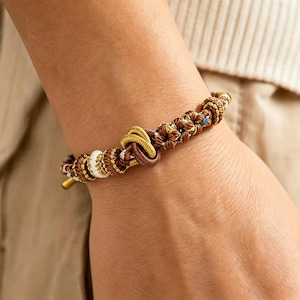 Può includere: Un bracciale in corda intrecciata con un nodo centrale color oro, affiancato da intricati motivi di fili marroni, bianchi e metallici. Il design incorpora diverse sezioni annodate e un terminale cilindrico liscio in metallo color oro.