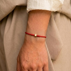 Può includere: Un braccialetto in cordino rosso caratterizzato da una singola piccola perlina cubica color oro centrata sul filo. Il braccialetto è realizzato con un sottile cordino rosso intrecciato che avvolge il polso, fissato con un nodo e un cubo dorato.