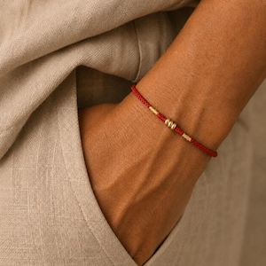 May include: A thin red cord bracelet featuring three small, smooth, metallic gold-toned beads centered along the string. The bracelet is constructed from a simple, continuous red thread that encircles the wrist, providing a minimalist aesthetic.