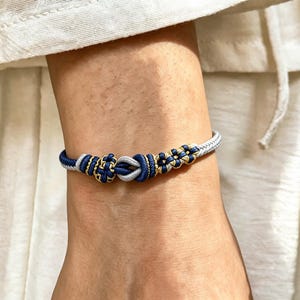 Pode incluir: Uma tornozeleira com um design de cordão trançado em azul-marinho, cinza-claro e dourado metálico. A pulseira apresenta um detalhe de nó central ladeado por nós decorativos menores em forma de flor, feitos de cordão sintético trançado.