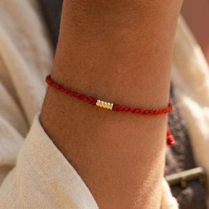 Puede incluir: Una pulsera fina de cordón rojo que presenta una pequeña cuenta cilíndrica de tono dorado con textura estriada en el centro. La pulsera está fabricada con un material textil trenzado y se asegura alrededor de la muñeca con un nudo simple.