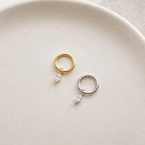 Puede incluir: Dos pendientes de aro, uno dorado y otro plateado, cada uno con una gema transparente en forma de lágrima colgante. Los pendientes se muestran sobre una superficie blanca ligeramente texturizada.
