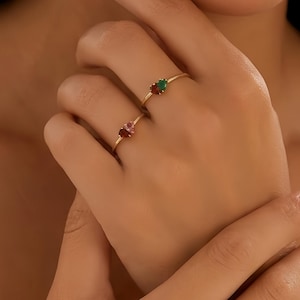 May include: Two gold rings, each with a gemstone. One ring features a red gemstone, and the other has a green gemstone. The rings are displayed on a hand, showcasing their delicate design and vibrant colors.