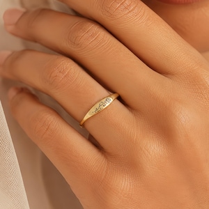 May include: A gold ring with a floral design. The ring is worn on a finger. The ring has a rectangular shape with rounded edges. The ring is a bright gold colour and has a delicate floral pattern engraved on the top.