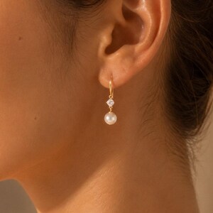 May include: A gold drop earring featuring a small, clear, round-cut gemstone set in a metal prong setting, suspended from a thin gold wire hook. Below the gemstone hangs a single, smooth, white spherical pearl, creating a delicate dangle design.