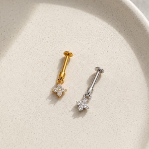 May include: Two small, dangling earrings. One is gold-toned, the other silver-toned. Each features a small, four-petal flower design with clear, sparkling stones. The earrings are displayed on a white, textured surface.
