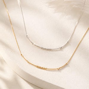 May include: Two delicate necklaces, one in gold tone and one in silver tone, each featuring a thin chain with a central curved bar element adorned with a series of small, round beads arranged in a line along the center of the curved bar.