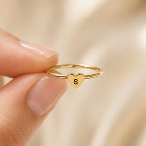 May include: A thin gold-toned ring featuring a small, flat heart-shaped charm centred on the band. The heart charm is engraved with the capital letter S in a clean, black sans-serif font. The ring has a polished, smooth metallic finish throughout.