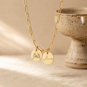 Puede incluir: Un collar de cadena tipo clip dorado con dos colgantes circulares. Un colgante muestra un grabado de líneas detallado de un conejo, mientras que el otro colgante tiene grabado el nombre Aria en una fuente de escritura cursiva.