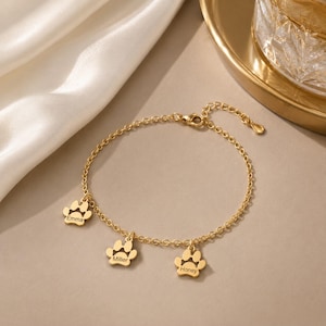 Puede incluir: Una pulsera de cadena en tono dorado con tres dijes colgantes en forma de huella. Cada dije está grabado con un nombre: Emma, Miller y Honey. La pulsera incluye una delicada cadena de eslabones y un pequeño cierre de mosquetón.