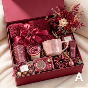 May include: A maroon gift box containing a chunky knit blanket with a satin ribbon, a floral arrangement, a pink speckled mug, a candle, a small speaker, herbal tea, hot cocoa, hand cream, a scrunchie, chocolates, and a small ornament.