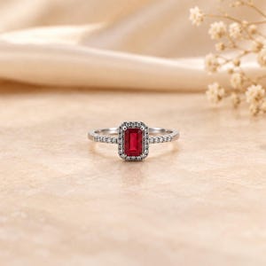 May include: A silver-toned ring featuring a rectangular, emerald-cut red gemstone at the center, surrounded by a halo of small, clear stones. The band is thin and also set with small, clear stones along the sides of the central setting.