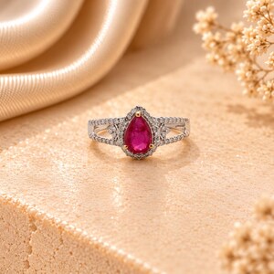 Puede incluir: Un anillo de tono plateado con una piedra preciosa central de color rosa vibrante en forma de pera, rodeada por un halo de pequeñas piedras claras. La banda se divide en dos brazos delicados, cada uno incrustado con diminutas piedras claras.