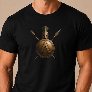 May include: A black short-sleeved t-shirt featuring a centered graphic of a bronze-colored Spartan helmet positioned above a circular shield with a large Greek letter Lambda, flanked by two crossed spears in a matching metallic bronze tone.