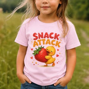 Könnte beinhalten: Ein hellrosa T-Shirt mit kurzen Ärmeln und einem Grafikdruck eines gelben Kükens, das eine große rote Erdbeere hält. Über dem Küken steht der Text SNACK ATTACK in fetten roten und orangefarbenen Buchstaben, umgeben von kleinen roten Herzen.