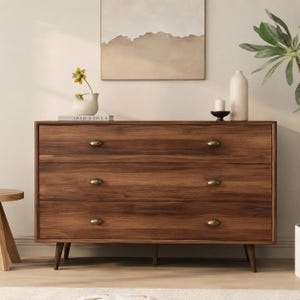 May include: A rectangular wooden dresser featuring three horizontal drawers with rounded brass-toned pulls. The furniture piece is supported by four tapered wooden legs and displays a rich brown wood grain finish throughout its exterior frame.