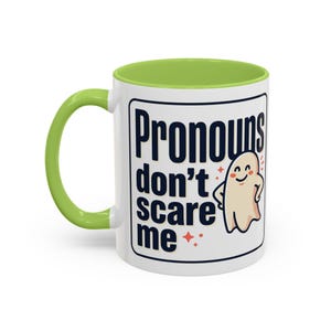 Op de afbeelding: Een witte keramische mok met een limoengroene binnenkant en handvat. De buitenkant heeft een zwarte rechthoekige rand met de donkerblauwe tekst "Pronouns don't scare me" naast een kleine, lachende, beige cartoongeest en rode sterren.