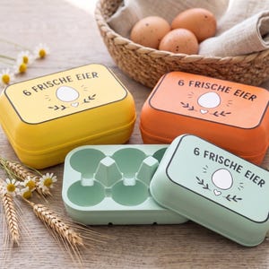 Könnte beinhalten: Drei rechteckige Eieraufbewahrungsboxen in Gelb, Orange und Hellgrün. Jeder Deckel ist mit dem Text 6 FRISCHE EIER, einer zentralen Eierillustration und Blattmotiven versehen. Eine Box ist geöffnet und zeigt das geformte Innere.