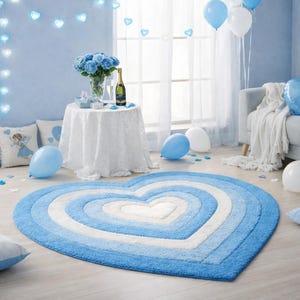 Puede incluir: Una alfombra en forma de corazón con capas concéntricas de azul y blanco. La alfombra está sobre un suelo de madera clara, con globos azules, una mesa con un mantel de encaje blanco y rosas azules en el fondo.