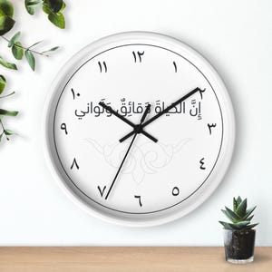 Op de afbeelding: Een ronde wandklok met een witte wijzerplaat, een witte rand en zwarte wijzers. De klok heeft zwarte Arabische cijfers van één tot twaalf in een cirkel. Arabische tekst staat in het midden boven een lichtgrijs, decoratief patroon.
