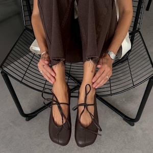 Può includere: Un paio di ballerine marrone scuro con punta arrotondata, una cucitura centrale verticale e sottili lacci abbinati che si annodano sul dorso del piede. Le scarpe hanno una finitura liscia e sono indossate con pantaloni marrone scuro.