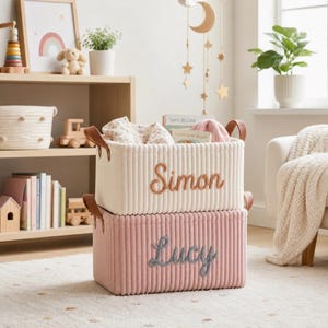 Op de afbeelding: Twee corduroy opbergmanden, één crème en één roze, op elkaar gestapeld. De crème mand heeft de naam "Simon" geborduurd in bruin, de roze "Lucy" in grijs. Beide hebben bruine leren handvatten.