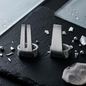 May include: Two silver-toned metal rings with a brushed finish. Each ring features a tall, vertical, tapered extension rising from the band. The left ring has a central vertical slit, while the right ring is solid. Both rings have engraved text.