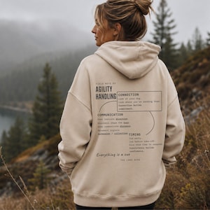 May include: A beige hooded sweatshirt featuring black text on the back. The print includes the words Agility Handling, Connection, Communication, and Timing, along with instructional phrases about dog training and the text Everything is a cue.