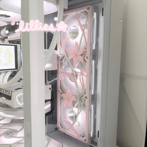 May include: A white computer case interior featuring a custom fan grill cover decorated with light pink lily flowers and green vines. The word "lillies" is written in a cursive white font with small star and flower icons in the upper left corner.