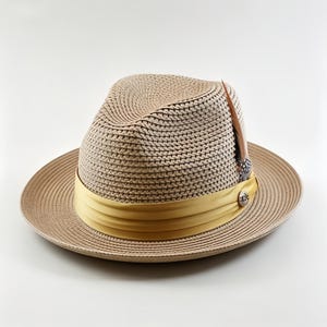 May include: A beige woven straw fedora hat featuring a wide, smooth mustard yellow ribbon band around the crown. A small, decorative silver-toned metal emblem is attached to the ribbon, and a single brown and white patterned feather is tucked in.