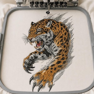 May include: An embroidery hoop holding off-white fabric with a detailed machine-embroidered design of a snarling leopard. The leopard is depicted in shades of orange, black, and white, with its claws extended forward and a fierce expression.