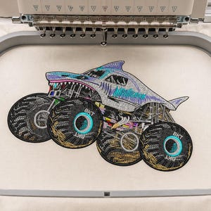 May include: An embroidery design of a shark-shaped monster truck featuring a silver body, sharp teeth, and large black tires with BKT branding, secured within a grey plastic embroidery hoop on a piece of off-white fabric under a machine needle.