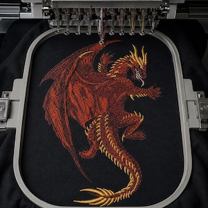 May include: An embroidery machine hoop holds black fabric featuring a detailed stitched design of a red dragon with gold accents. The dragon is depicted in a dynamic pose with wings spread, sharp claws, and a long tail, positioned under the machine needles.