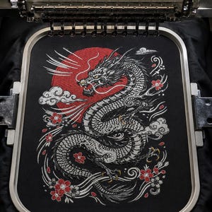 May include: An embroidery hoop holding black fabric with a detailed machine-stitched design of a long, winding dragon in white and grey thread. The dragon is positioned in front of a large, circular red sun, surrounded by white clouds and red flowers.