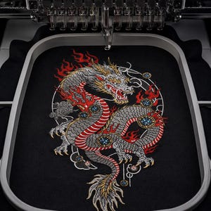 May include: An embroidery machine hoop containing black fabric with a detailed, multi-colored dragon design. The dragon features silver scales, red flames, and gold accents, surrounded by a thin white circular outline and small circular motifs.