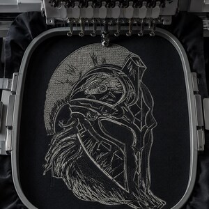 Peut inclure: Une pièce de tissu noir fixée dans un tambour à broder présentant un motif en fil blanc détaillé représentant un casque spartiate. Le casque est représenté de profil avec des lignes et des ombres complexes, sous une tête de machine à broder.