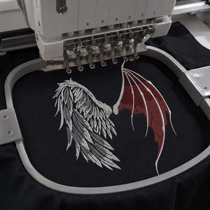 以下が含まれることがあります： 黒い布に刺繍を施す刺繍機。デザインは対照的な2つの翼で構成されており、左側には詳細な白い羽の翼、右側には暗い赤色の尖ったコウモリのような翼が描かれ、刺繍枠の中央に配置されています。