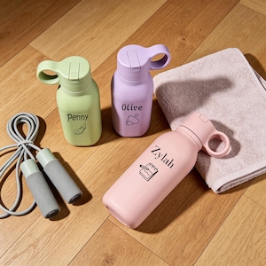 Op de afbeelding: Drie waterflessen in lichtgroen, lavendel en lichtroze, elk met een naam en een klein icoontje. De groene fles toont Penny met een aubergine, de lavendelkleurige Olive met wolken, en de roze fles Zylah met een notitieblok. Een springtouw en een handdoek zijn inbegrepen.