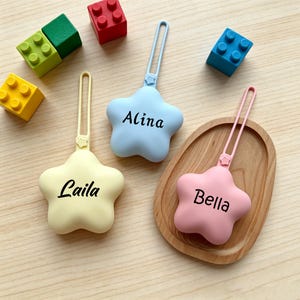 May include: Three star-shaped silicone bag tags with loops, each featuring a custom name in black script: Laila on a yellow tag, Alina on a light blue tag, and Bella on a pink tag. The tags are arranged alongside colorful plastic building blocks.