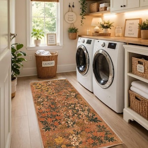 May include: A rectangular runner rug featuring a warm orange background with a dense pattern of small white, yellow, and orange flowers interspersed with dark green leaves, positioned on a light wood floor in a laundry room with two white appliances.