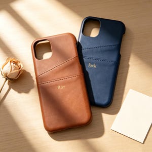 May include: Two phone cases with card slots, one in tan leather with the name Ray embossed in gold, and one in navy blue leather with the name Jack embossed in gold. Both cases feature a diagonal pocket design and a large camera cutout.