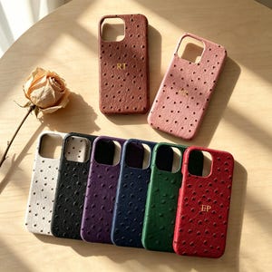 Puede incluir: Una colección de ocho fundas para smartphone con un patrón texturizado de piel de avestruz en varios colores, incluyendo marrón, rosa, blanco, negro, morado, azul marino, verde y rojo. Algunas fundas están personalizadas con iniciales doradas.